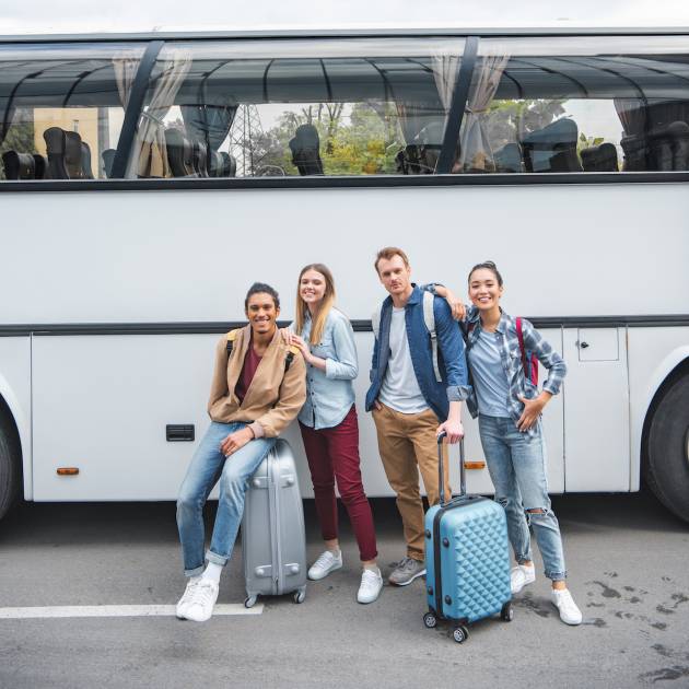 young multicultural friends with wheeled bags posing near travel bus at street