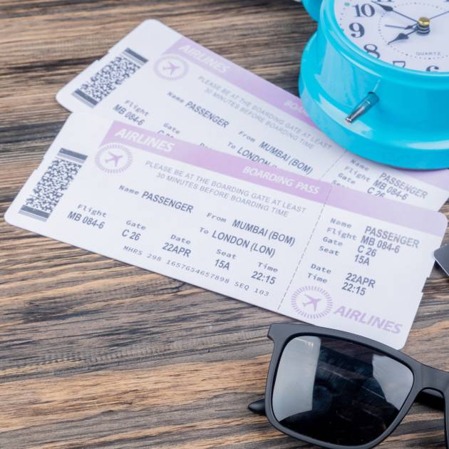 top view of plane tickets with blue alarm clock sunhat and sunglasses on a wooden background